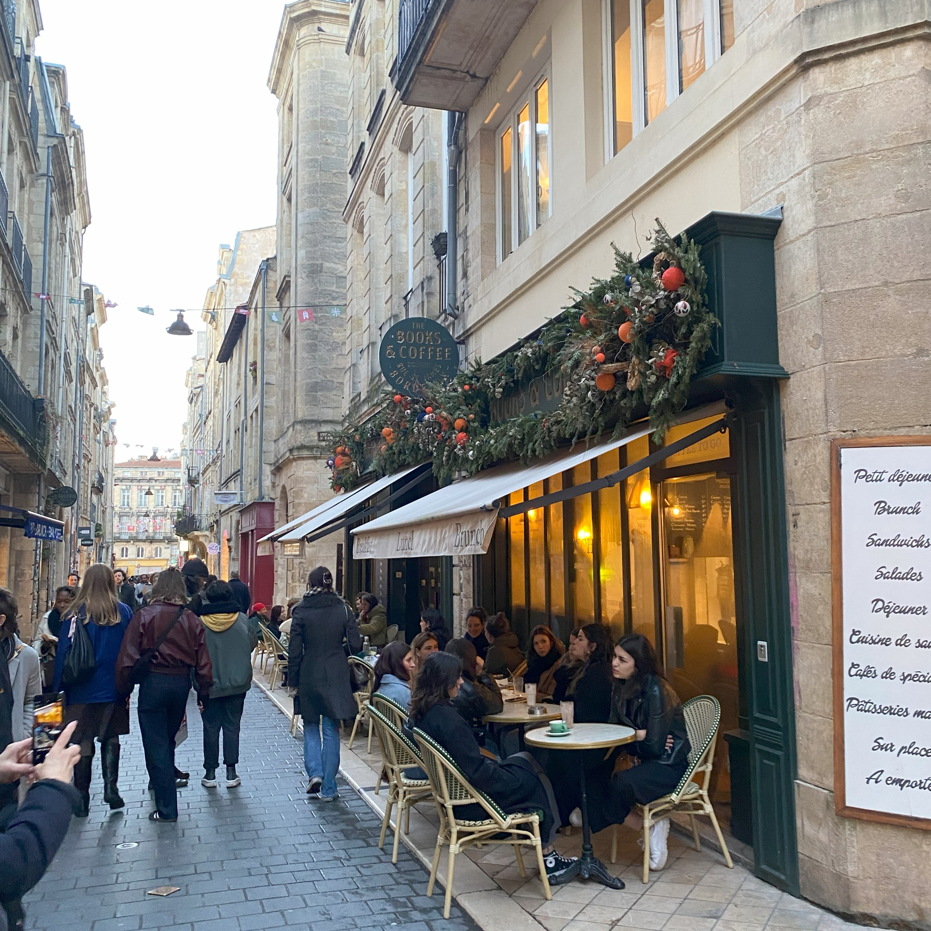 Pessoas em uma rua de paralelepípedos em uma cidade francesa. Mesas de café com clientes ocupam a calçada, enquanto pedestres caminham ao fundo. A fachada de uma cafeteria, decorada com guirlandas e luzes, se destaca à direita.